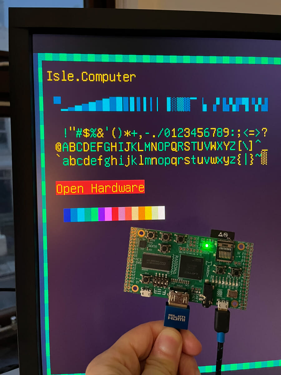 ULX3S FPGA dev board connected to widescreen monitor over HDMI cable showing text mode using internal system ROM. Text shows 'Isle.Computer' and basic Latin and block glyphs.